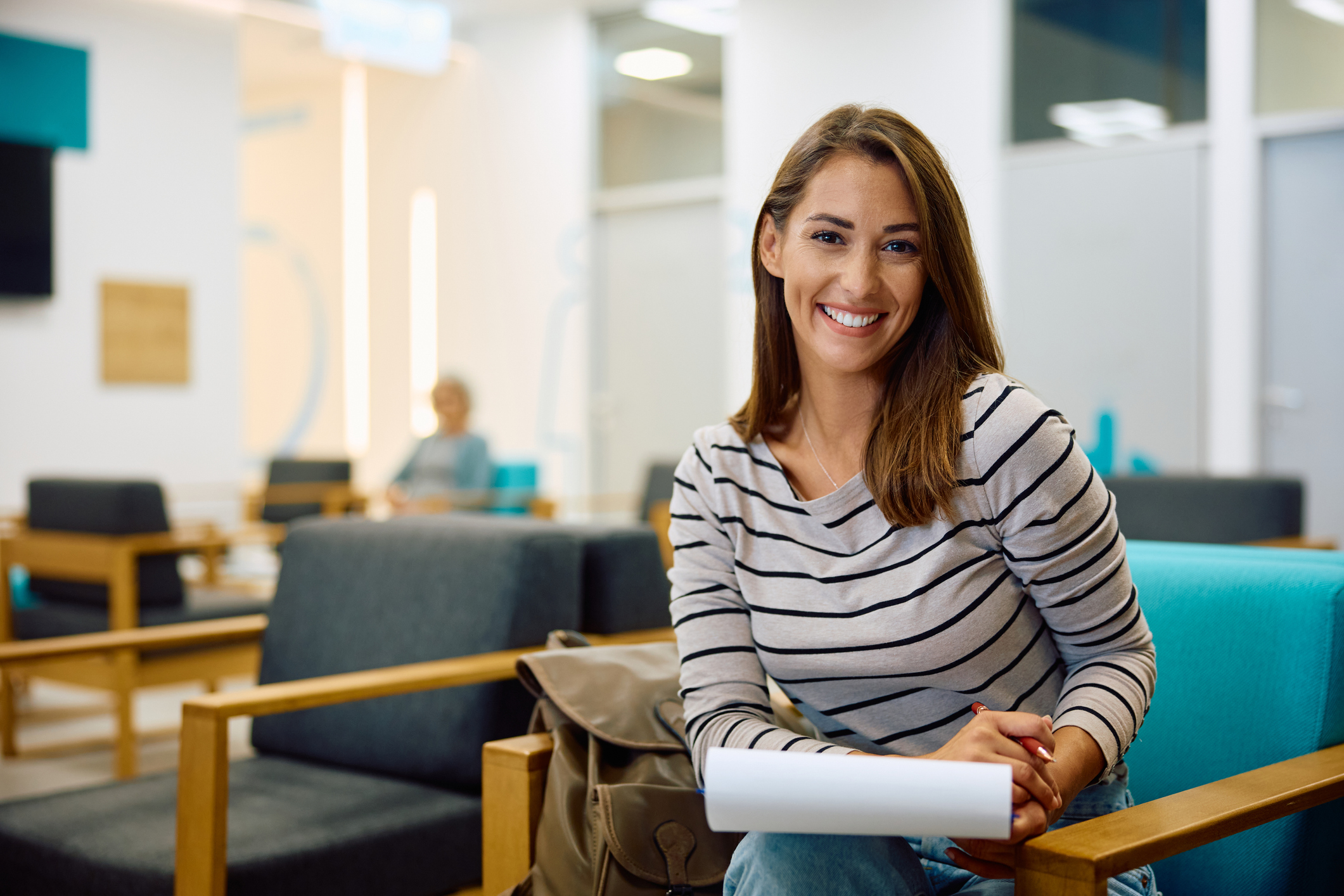 Happy woman filling out medical paperwork in waiting room at doctor’s office and looking at camera. Young happy woman writing her data on medical documents while waiting for doctor's appointment at the clinic and looking at camera.